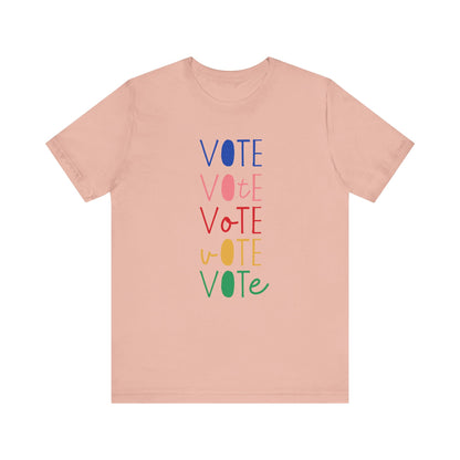 A peach short-sleeve t-shirt with a vertical graphic on the chest spelling 'VOTE' in alternating bright colors (blue, pink, red, yellow, green) with oval letterforms, shown on a plain white background.