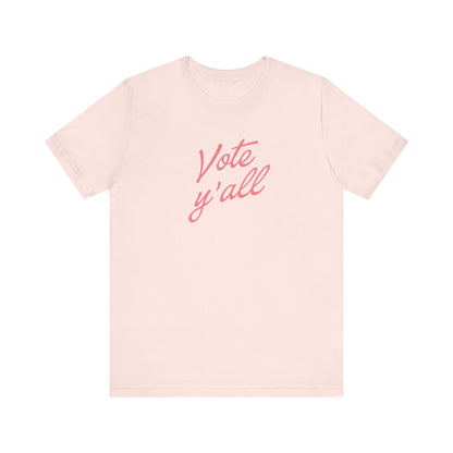 A light pink short-sleeve crewneck t-shirt featuring the phrase 'Vote y'all' printed in a pink cursive font across the chest, displayed on a clean white background.