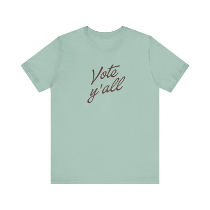 A sage green short-sleeve t-shirt with the phrase 'Vote y'all' printed in brown cursive script across the chest, displayed on a clean white background.