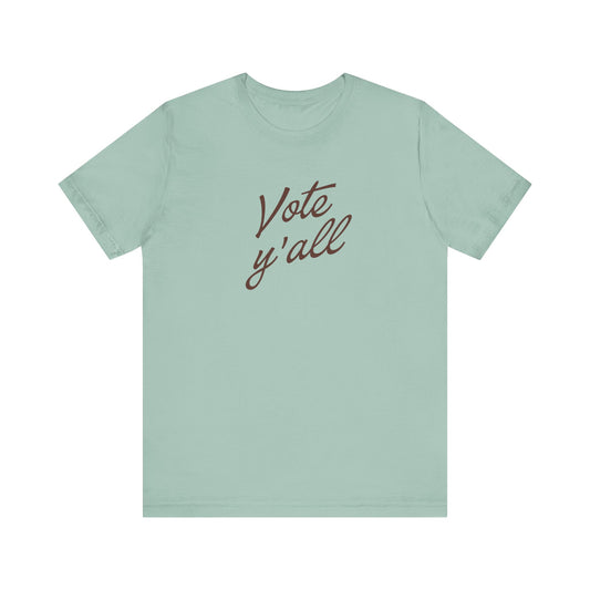 A sage green short-sleeve t-shirt with the phrase 'Vote y'all' printed in brown cursive script across the chest, displayed on a clean white background.