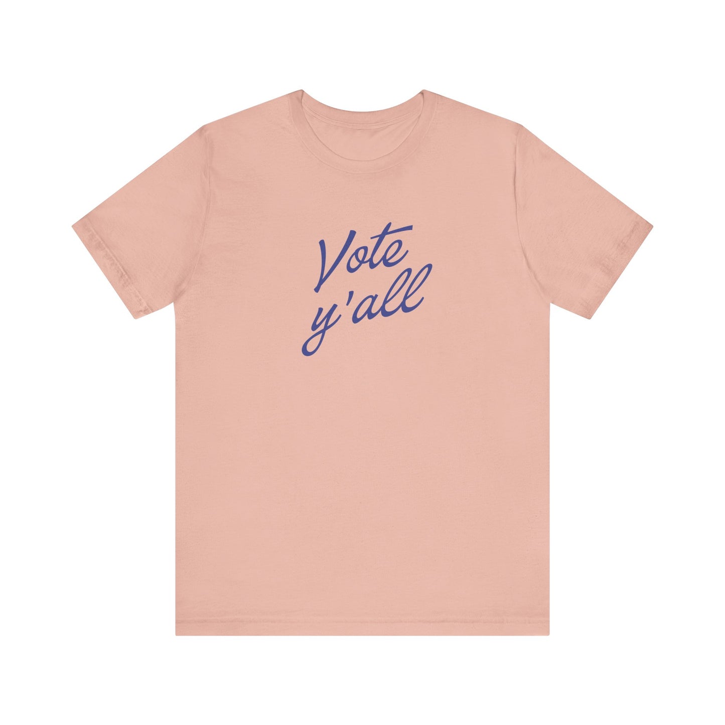 A light peach-pink short-sleeve t-shirt with the words 'Vote y'all' printed in a blue cursive script across the chest, displayed on a solid white background.
