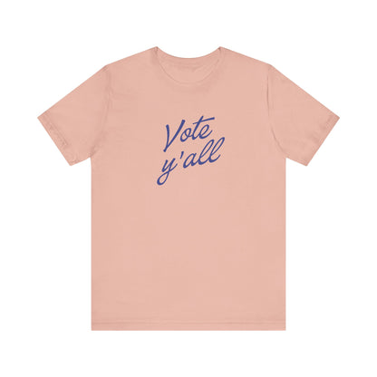 A light peach-pink short-sleeve t-shirt with the words 'Vote y'all' printed in a blue cursive script across the chest, displayed on a solid white background.