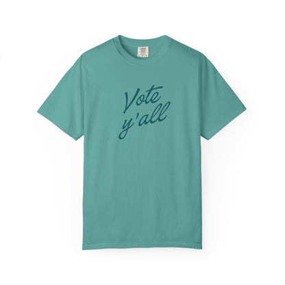 A short-sleeve crewneck t-shirt in a teal blue color, featuring the phrase 'Vote y'all' printed in a darker teal cursive script across the chest, displayed on a clean white background.