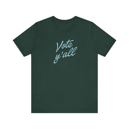 A dark green short-sleeve crewneck t-shirt featuring the phrase 'Vote y'all' printed in a light blue cursive font across the chest, displayed on a clean white background.
