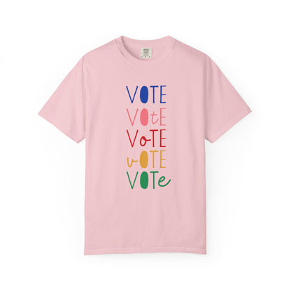 A light pink short-sleeve t-shirt with a vertical graphic spelling 'VOTE' in alternating blue, pink, red, yellow, and green colors, displayed on a clean white background.
