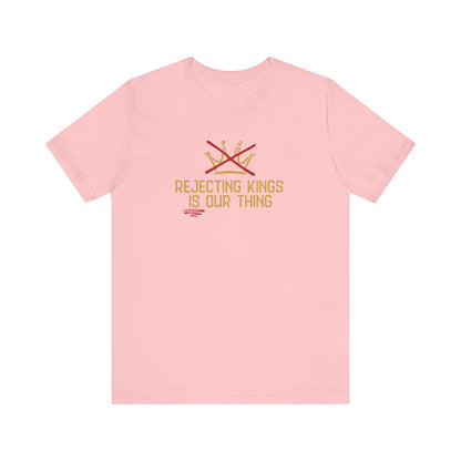 Pink t-shirt with a gold graphic sketch of a brown with a red X over it and gold words underneath "Rejecting Kings Is Our Thing" 
