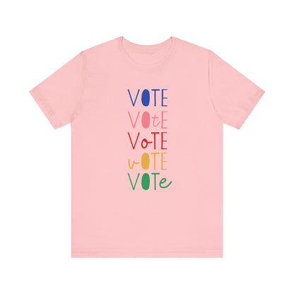 A light pink short-sleeve t-shirt with a vertical graphic on the chest spelling 'VOTE' in alternating bright colors (blue, pink, red, yellow, green) with oval letterforms, shown on a plain white background.
