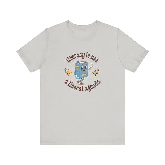 Light gray t-shirt with a graphic of a retro book character and "literacy is not a liberal agenda" brown text