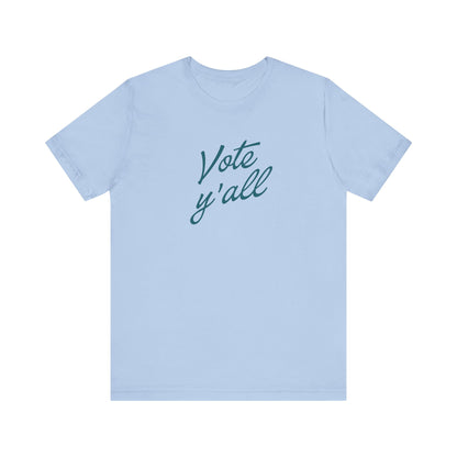 A light blue short-sleeve crewneck t-shirt featuring the phrase 'Vote y'all' printed in a dark teal cursive font across the chest, displayed on a clean white background.