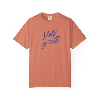 A short-sleeve crewneck t-shirt in a muted terracotta color, featuring the phrase 'Vote y'all' printed in blue cursive script across the chest, displayed on a clean white background.