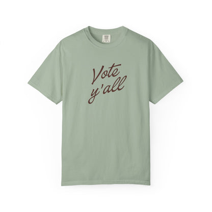 A short-sleeve crewneck t-shirt in a light green color, featuring the phrase 'Vote y'all' printed in brown cursive script across the chest, displayed on a clean white background.