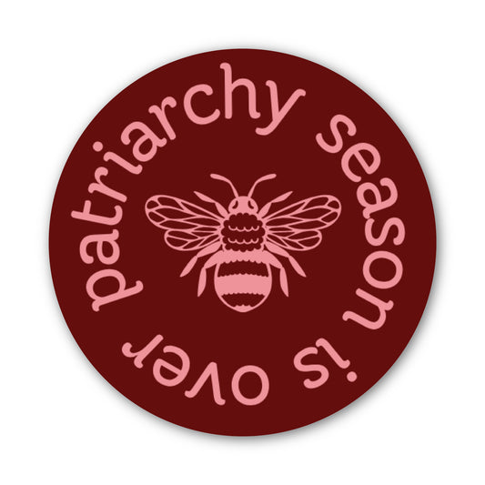 A round sticker or decal with a dark red background, featuring a pink bee in the center and the text 'patriarchy season is over' in pink around the edge.
