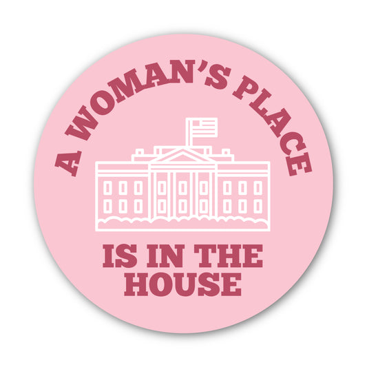 Light pink round sticker with words around the edge in dark pink "A Woman's Place Is in the House" with a white sketch graphic of the White House in the center.